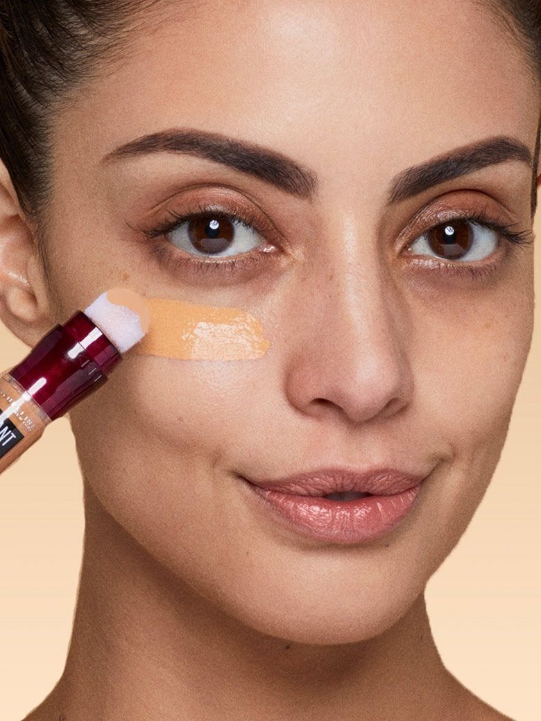 Mulher a aplicar o corretor de olheiras maybelline em fundo bege